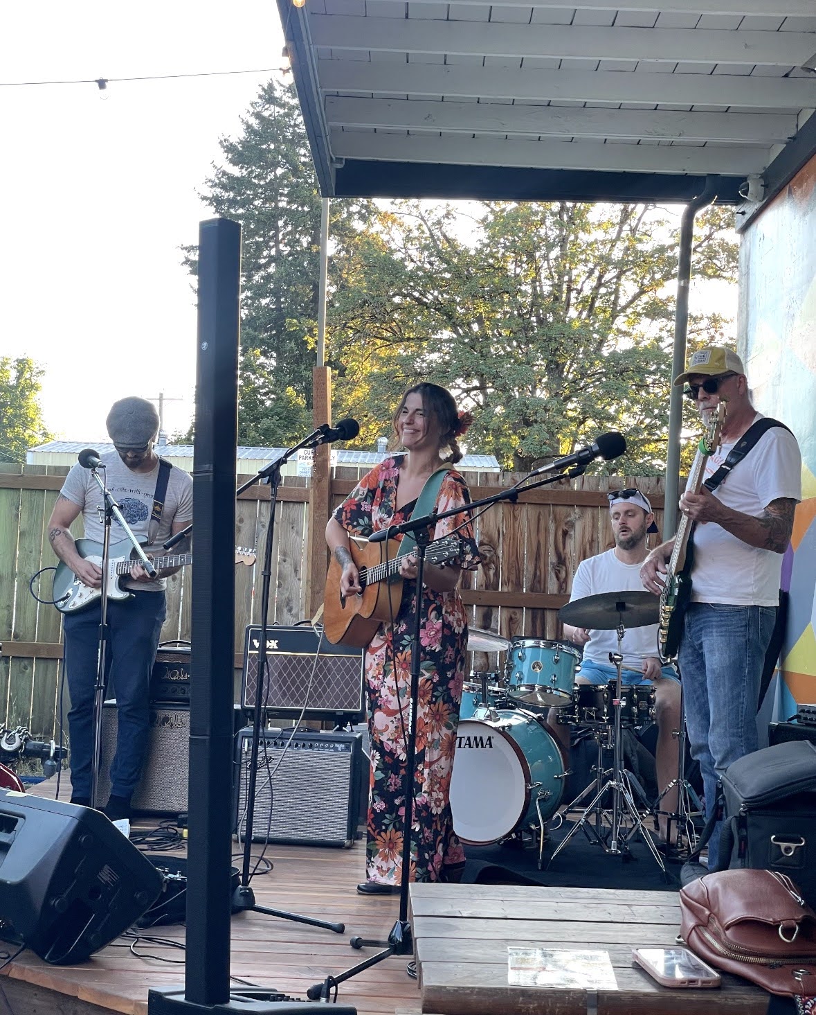 Big Fir playing outdoors at golden hour, the singer smiling mid-song with acoustic guitar while the band plays behind her