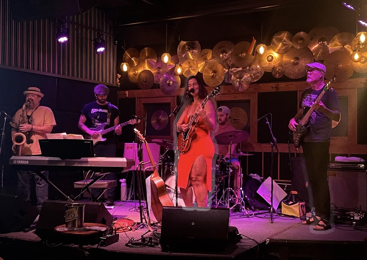 Big Fir on an indoor stage under warm and violet lights, singer in a red dress at the microphone with the full band behind her
