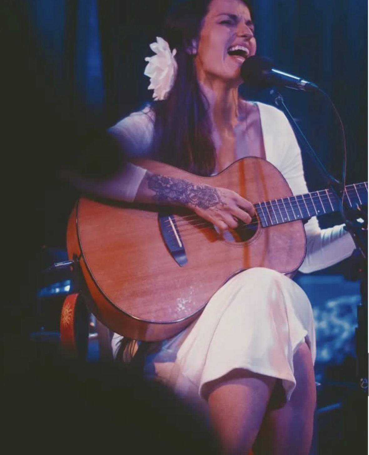 Sarah singing into a microphone while playing an acoustic guitar under blue stage light, a white flower in her hair