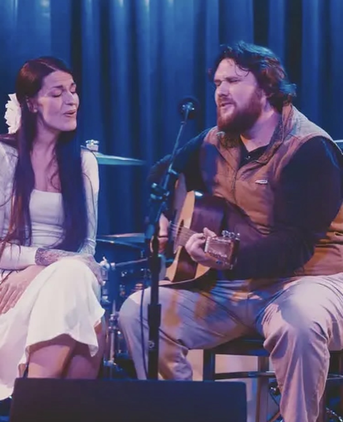 Two Big Fir musicians seated on stage singing together, one playing an acoustic guitar in front of a blue curtain