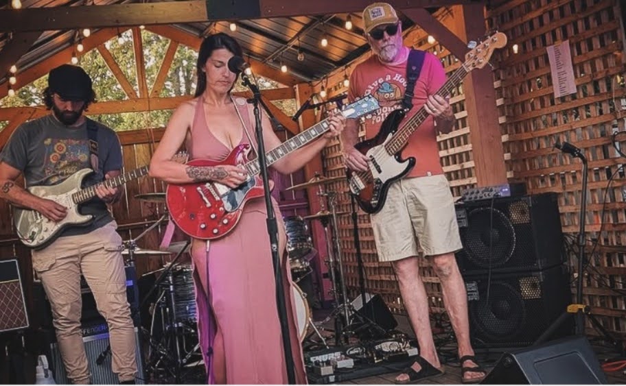 Big Fir on an outdoor covered stage: guitarist at left, singer in a pink dress playing a red hollow-body guitar, bassist at right